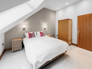 A bedroom with a sloped ceiling, a bed with white bedding, two bedside lamps, a wooden wardrobe, and a small bedside table. - Coach House - UK52274 (Helmsley)