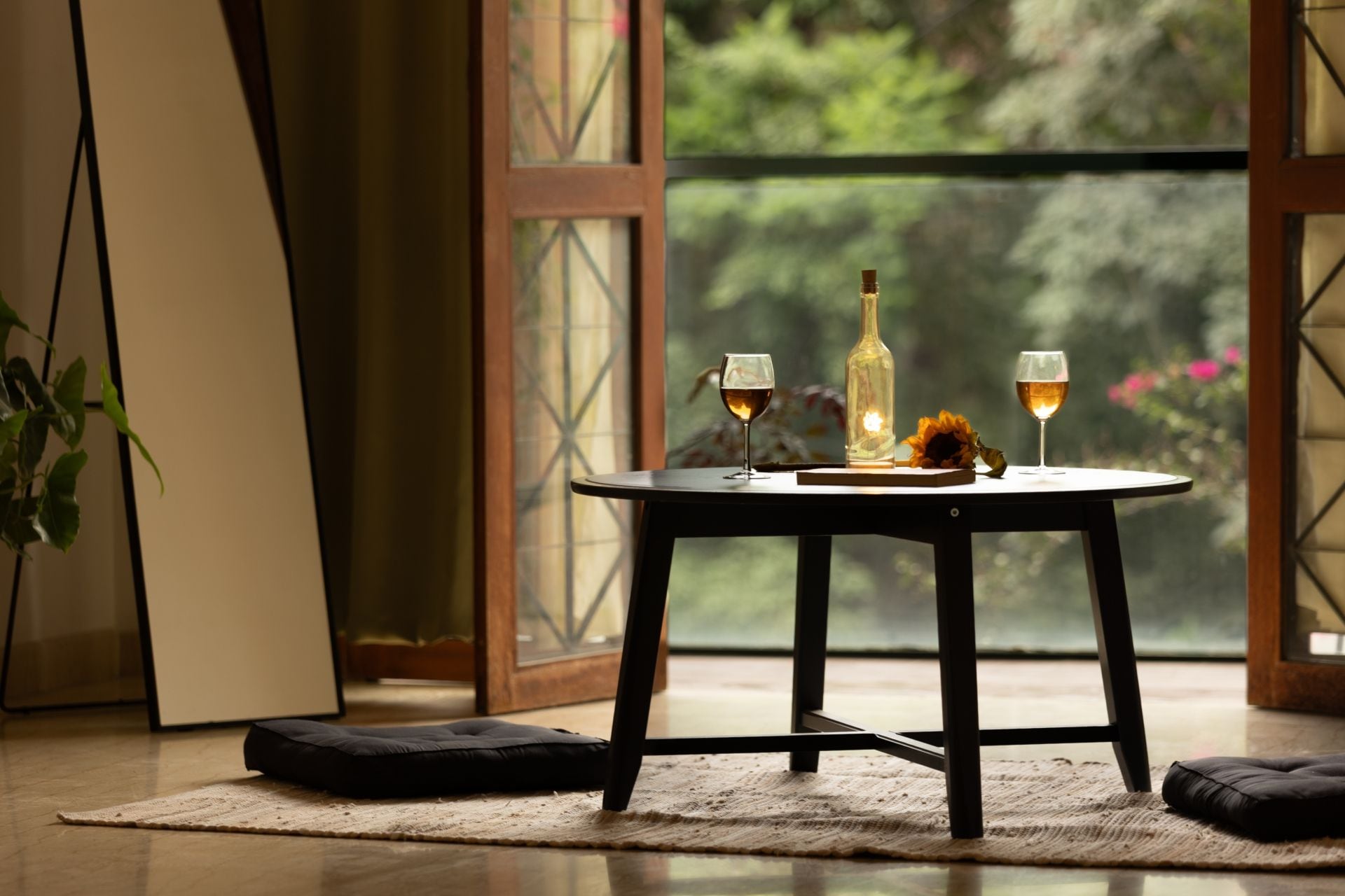 A round table with two glasses of wine, a bottle, and a sunflower on a rug.