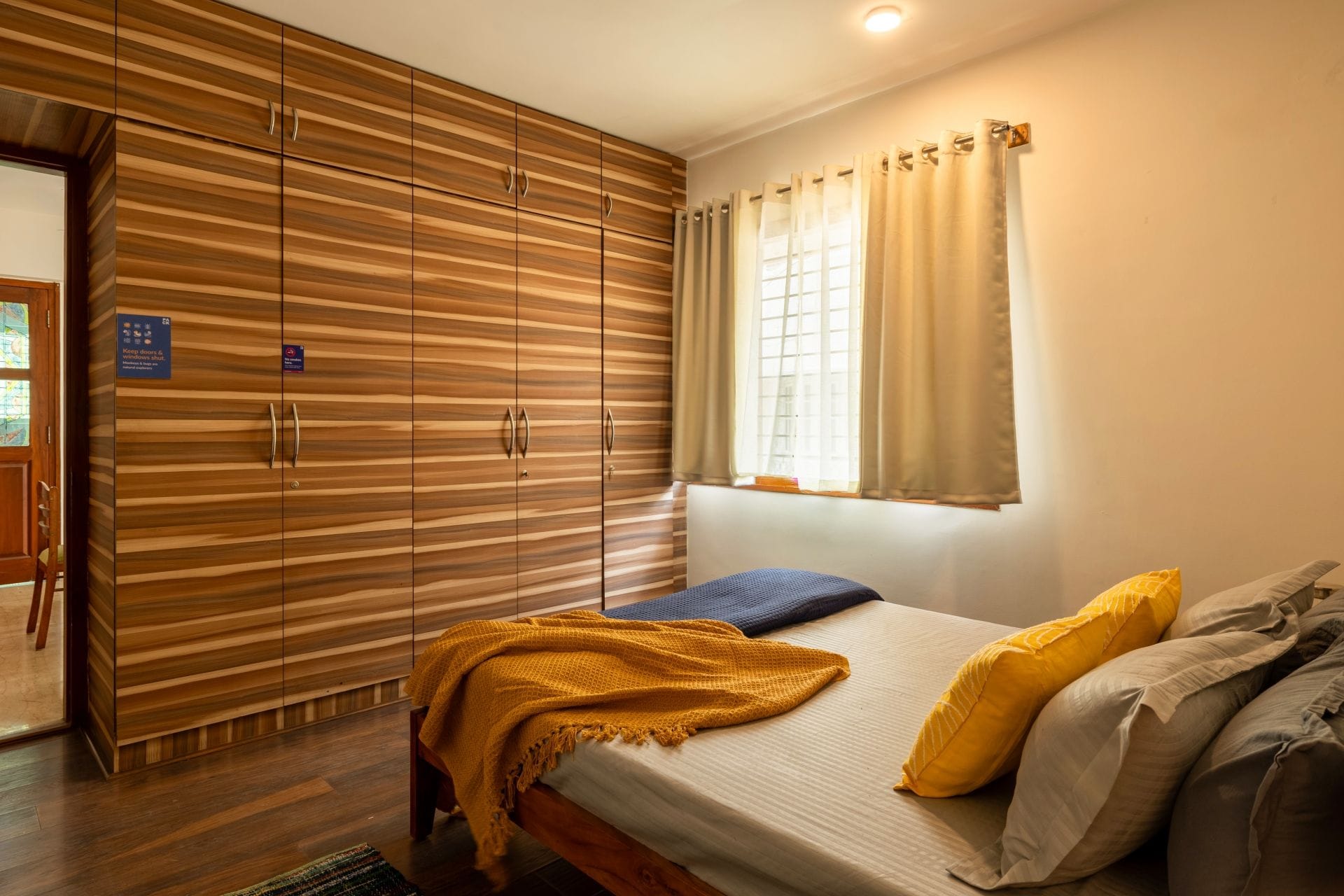 A bedroom with a bed, a wooden wardrobe, a window with curtains, and a door with a sign.