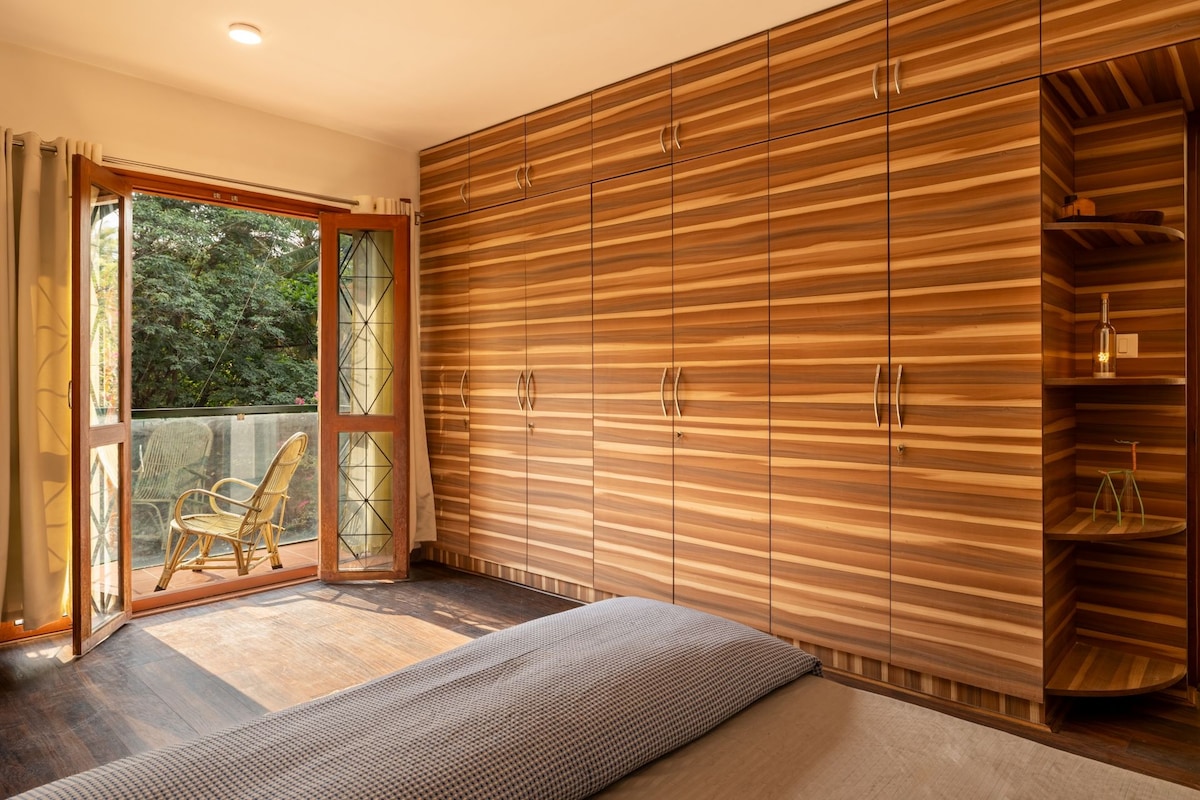 A room with a large wooden wardrobe, a glass door leading to an outdoor area with a chair, and a bed with a patterned bedspread.