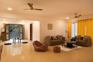 A spacious living room with two sofas, a coffee table, and a bean bag. There are ceiling fans, a window with yellow curtains, and a door leading to another room. - LiveAway Bengaluru (HSR) by PACK (Bengaluru)