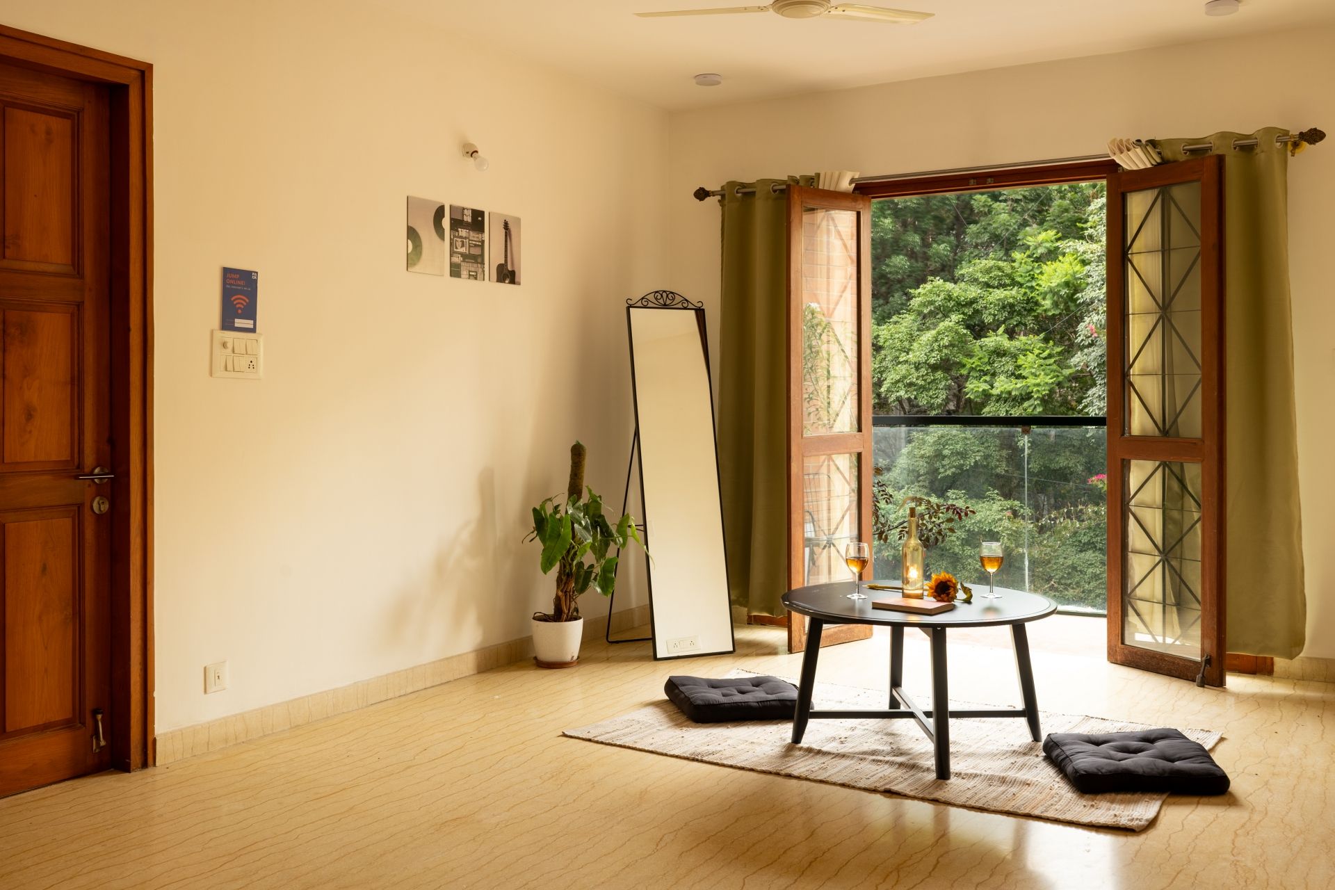 A room with a wooden door, a standing mirror, a potted plant, a round table with a cushion, and a view of greenery through a window.
