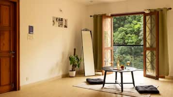 Habitación con puerta de madera, un espejo de pie, una planta en maceta, una mesa redonda con cojín y vista a follaje por una ventana.