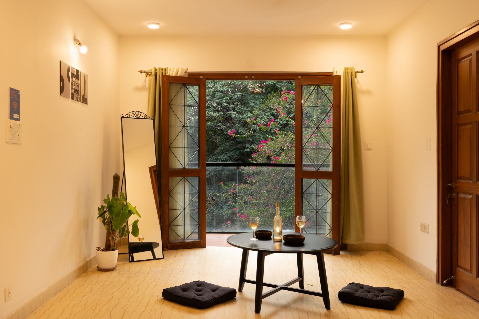 Una habitación de esquina con un ventanal grande, una mesita con dos cuencos y dos vasos, una planta en maceta y un cojín negro en el suelo.