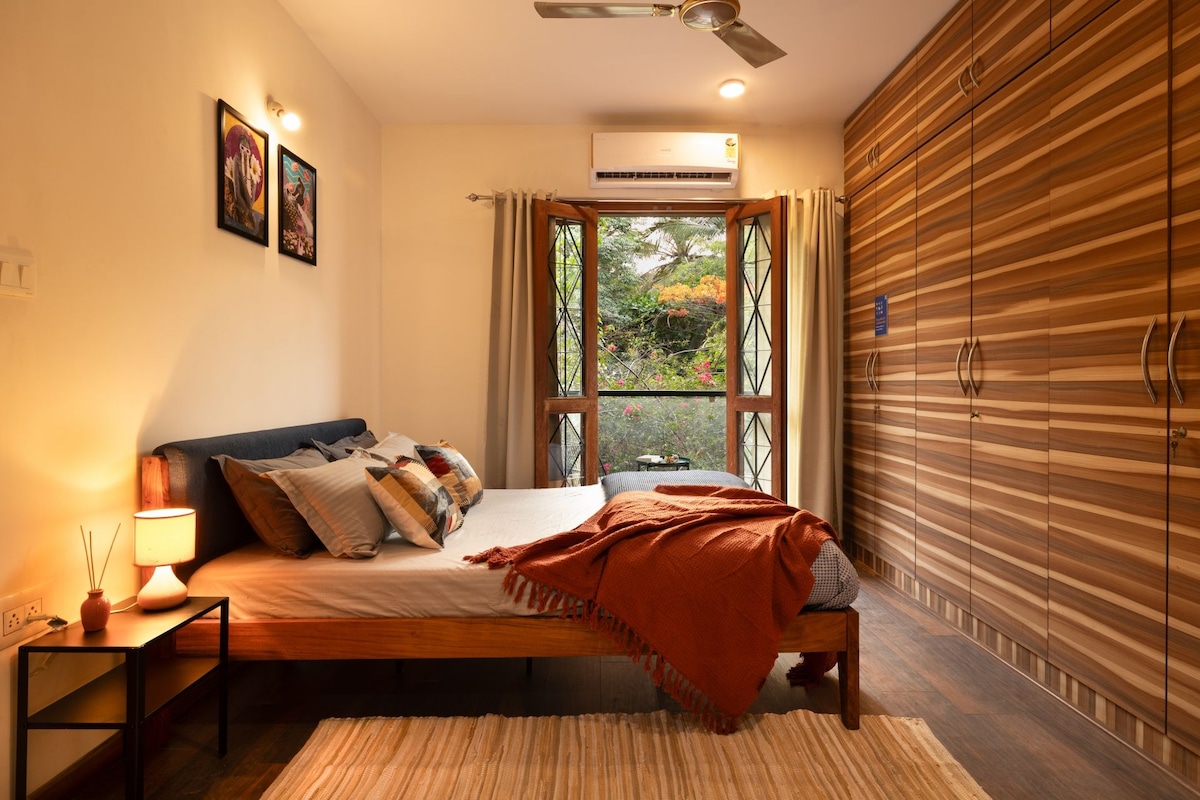 A bedroom with a bed, bedside table, lamp, and a view through a window.