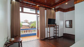 Quarto de hotel com soalho de madeira, televisão sobre suporte, cama e varanda com cortinas.