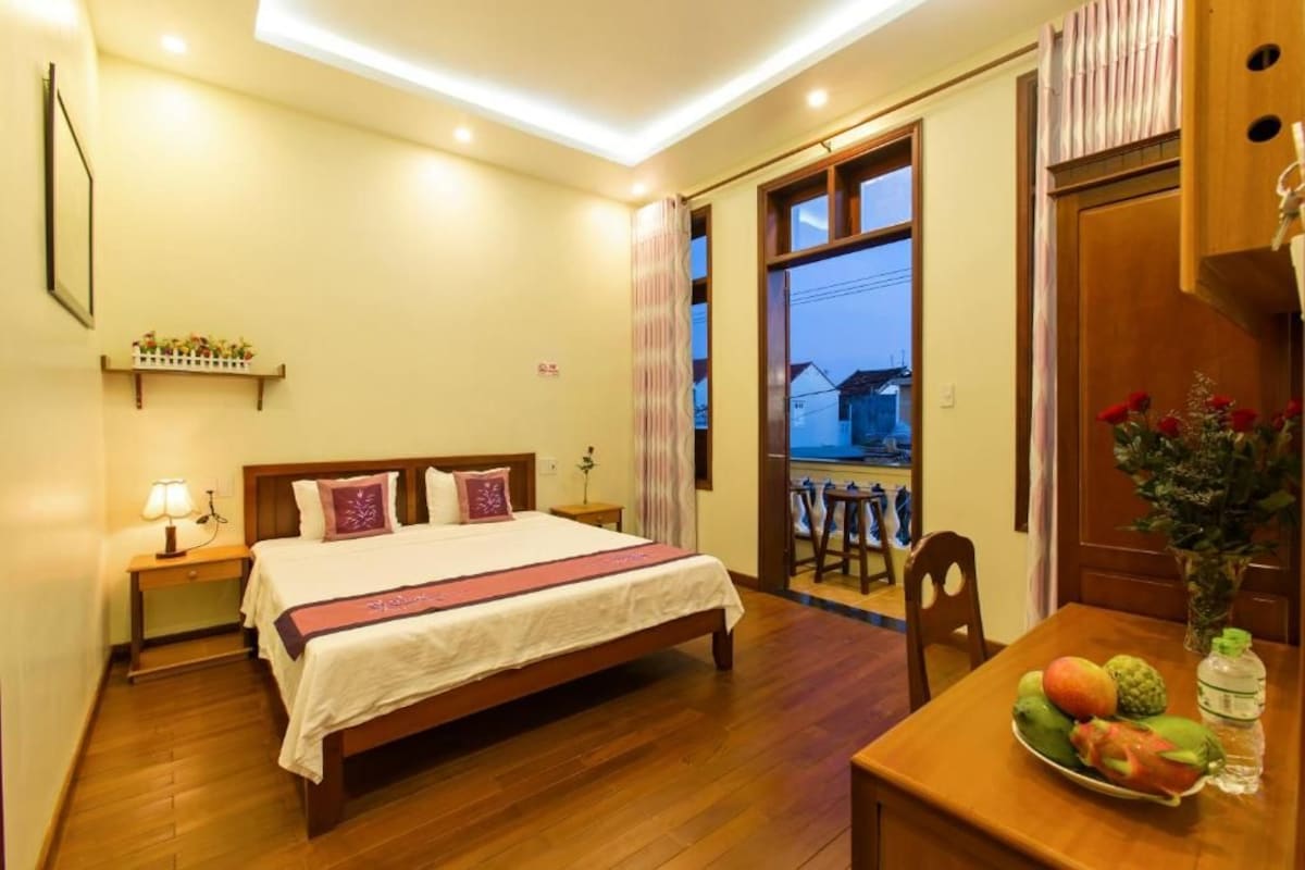 A hotel room with a bed, bedside table, lamp, and a dining table with fruit.
