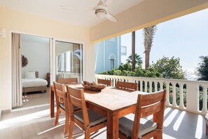 Ein Balkon mit Holztisch und Stühlen, ein Bett mit weißer Matratze und Blick auf Palmen und ein Gebäude.