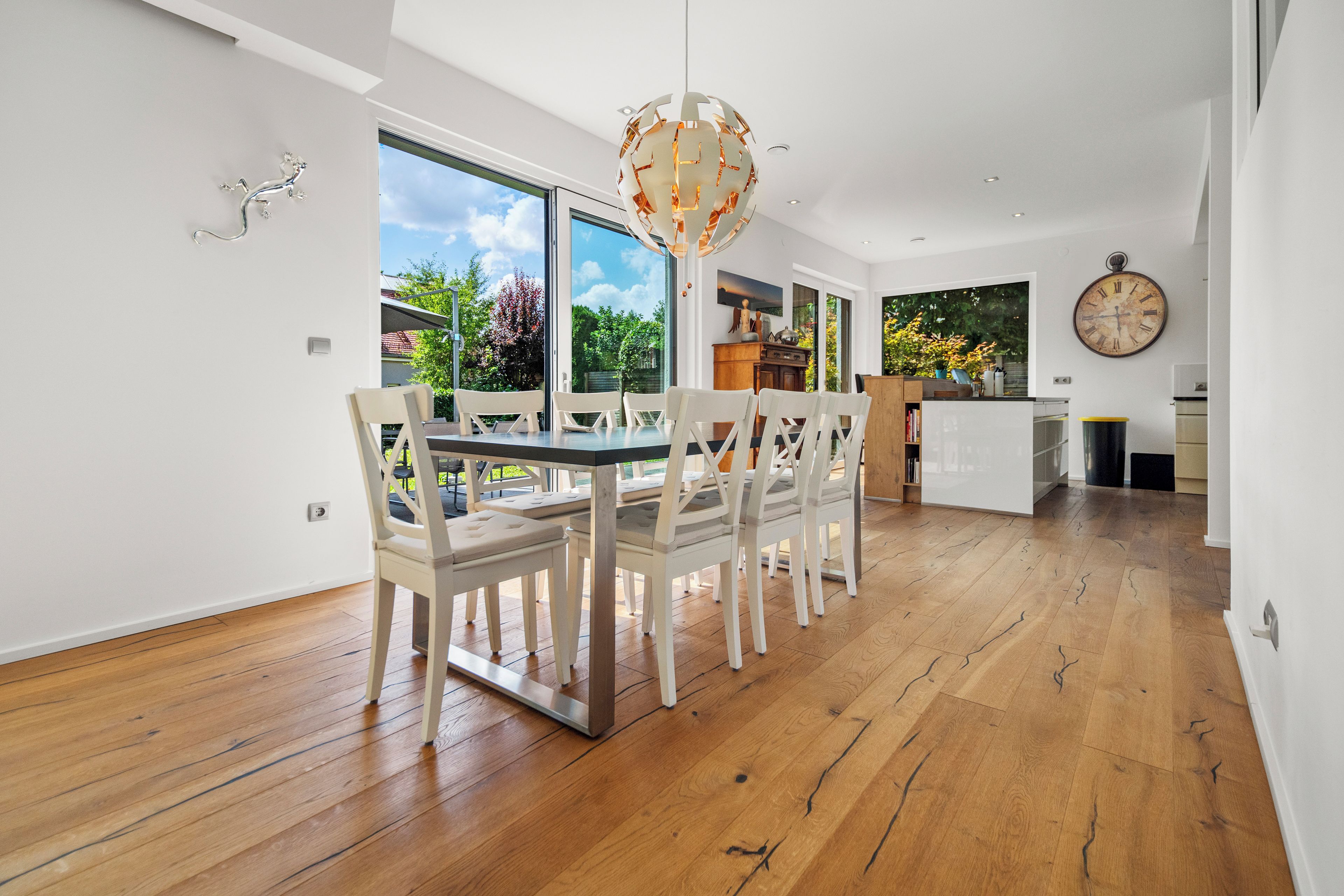 A spacious dining area with a modern chandelier, wooden flooring, and large windows offering a view of the outdoors.