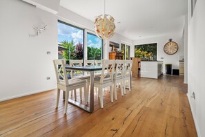 A spacious dining area with a modern chandelier, wooden flooring, and large windows offering a view of the outdoors.