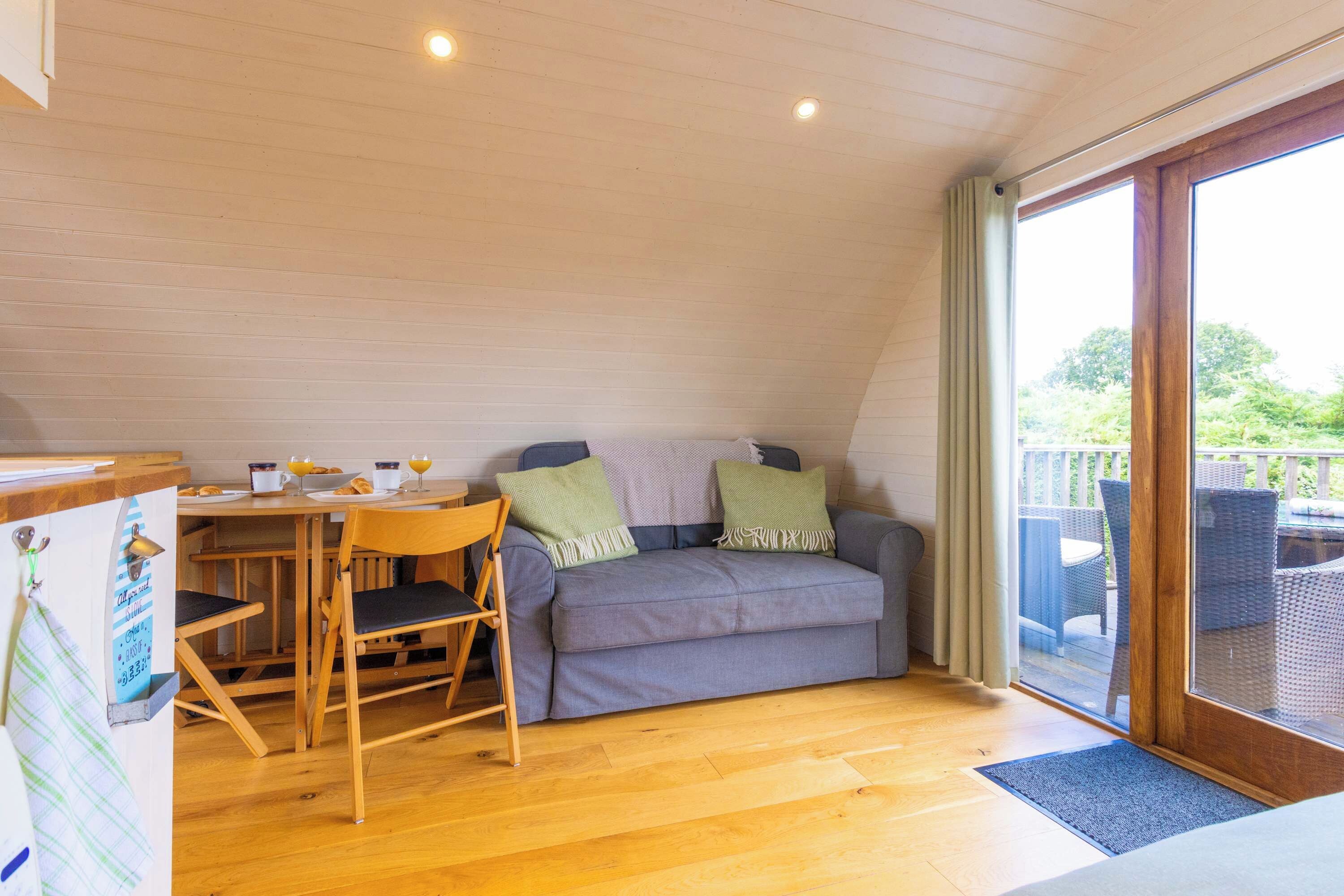 Ein gemütliches Zimmer mit Holzboden, einem grauen Sofa mit grünen Kissen, einem Holzesstisch mit Stühlen und einer großen Schiebeglasdoor zur Außenbereits.