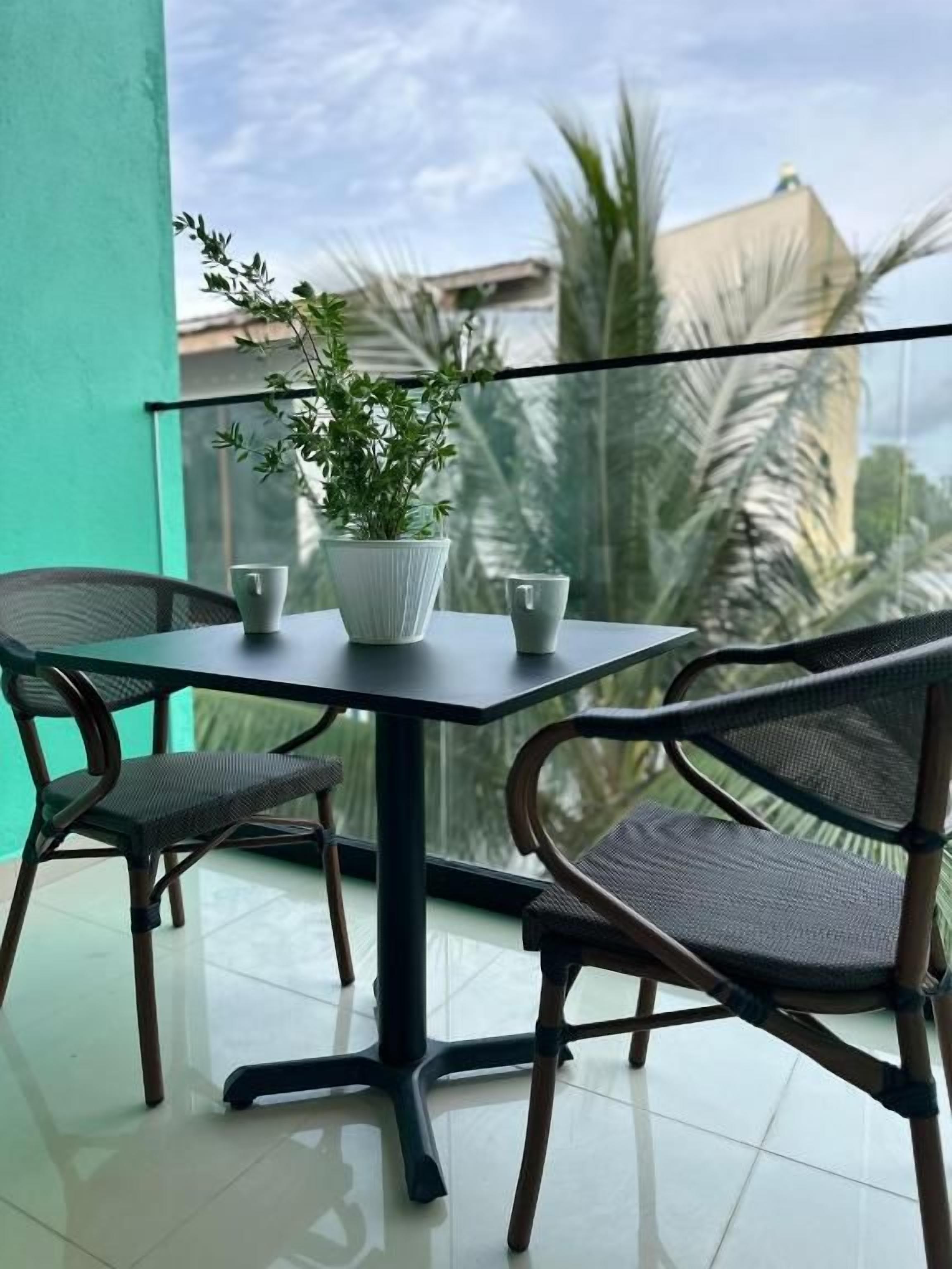 En balkon med et bord og stole, en potteplante og udsigt til palmer og bygninger.