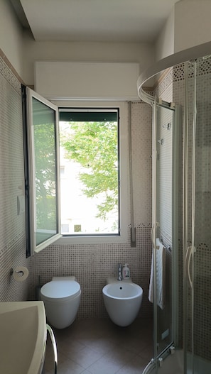 Une salle de bain avec deux lavabos, des toilettes, une douche et une fenêtre donnant sur un espace verdoyant.