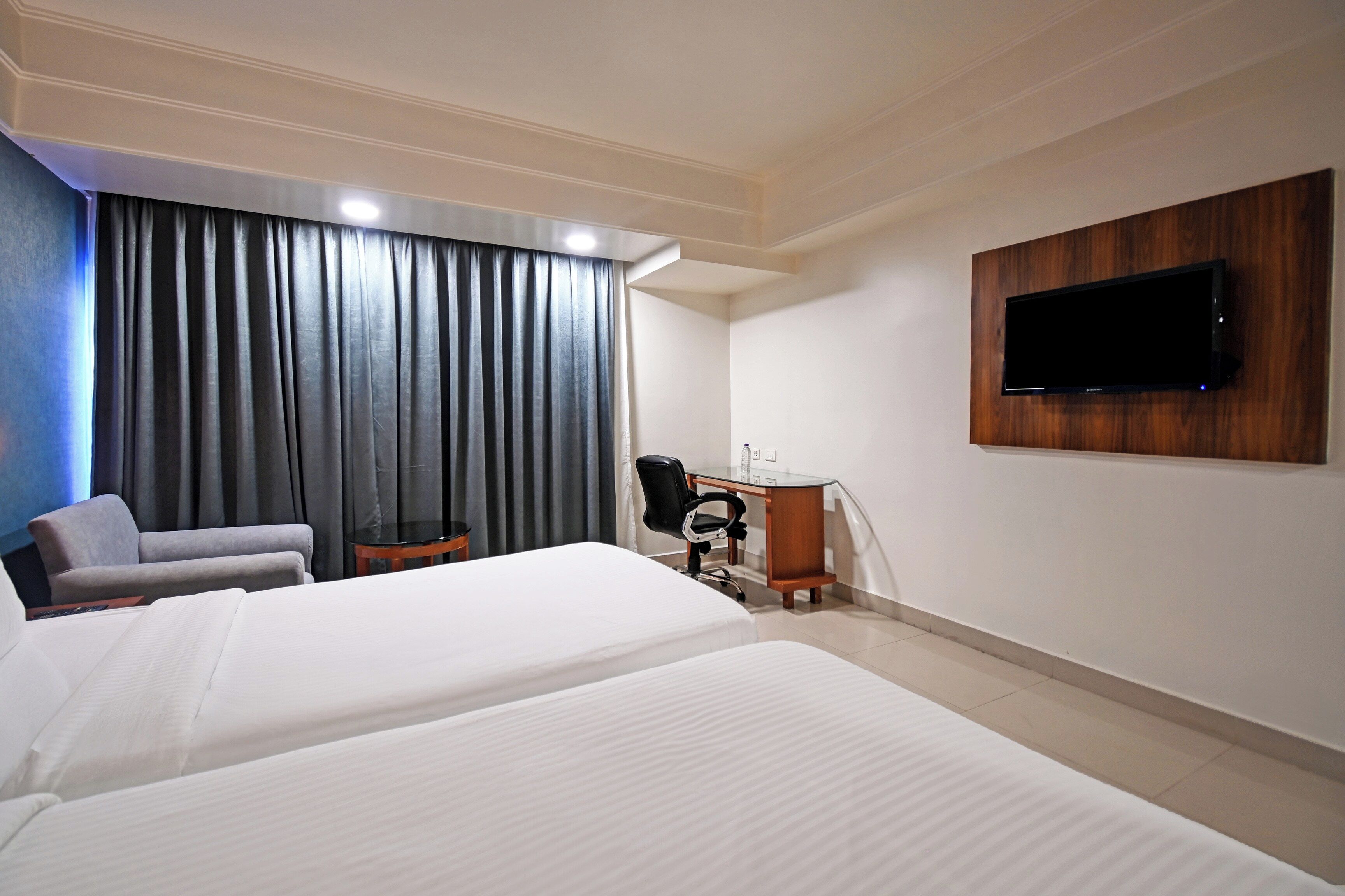 Habitación de hotel con dos camas, un escritorio, una silla, un televisor montado en la pared y un ventanal con cortinas.