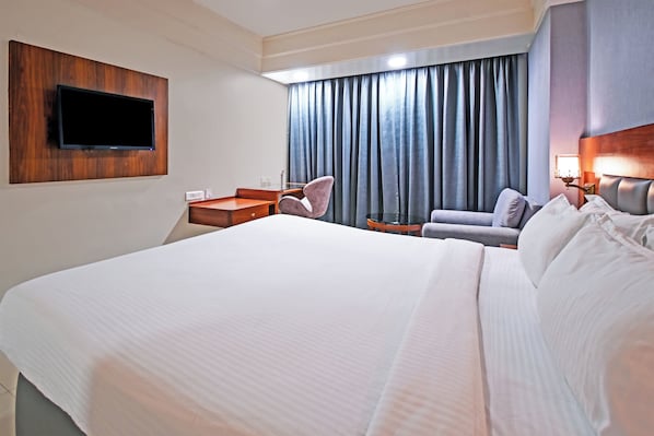 A hotel room with a large bed, a flat-screen TV mounted on the wall, a seating area with a sofa and armchairs, and a window with curtains.