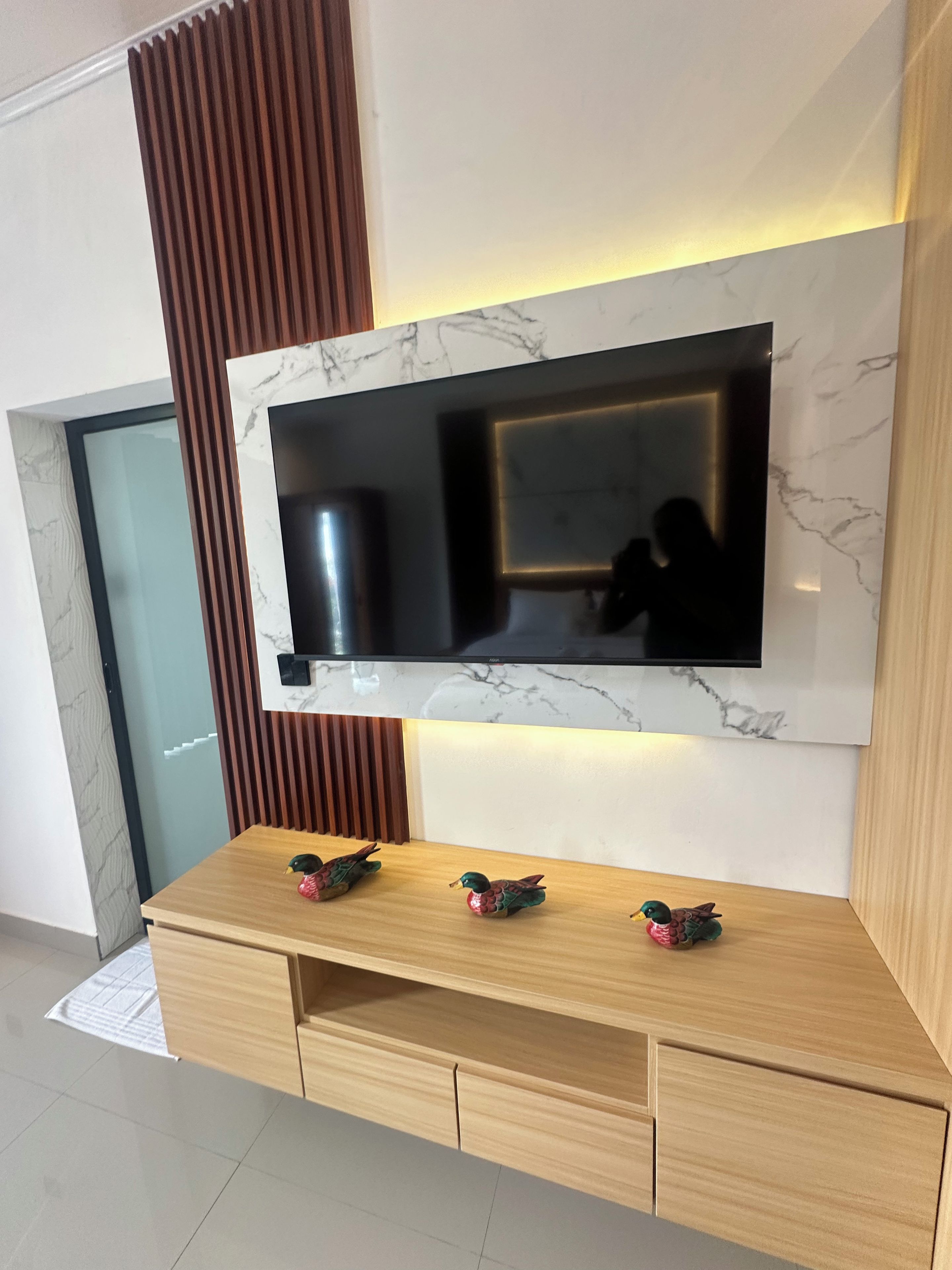 Una sala moderna con un televisor de pantalla plana montado en una pared de mármol, paneles de madera y un mueble bajo de madera con figuritas de patos decorativos.