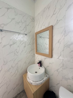 A bathroom with a marble wall, a wooden cabinet, a white sink with a black faucet, and a wooden-framed mirror. - Monte Batur (Bangli)