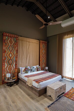 A bedroom with a large bed, a ceiling fan, a window with curtains, and a nightstand with a lamp. - ELIVAAS Prive Sravasti Forest View (Bengaluru)