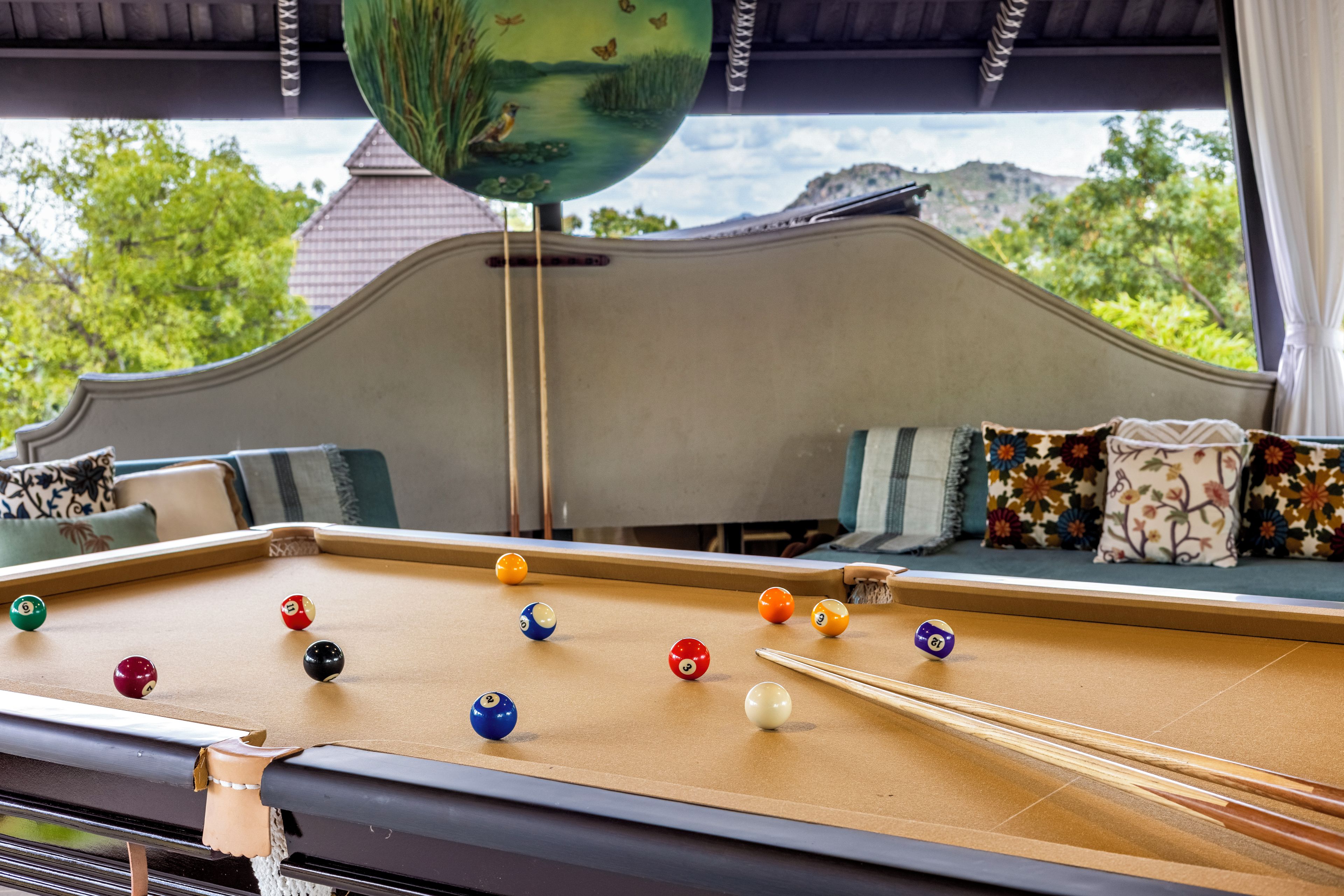A pool table with balls, a sofa with patterned cushions, and a view of greenery outside.