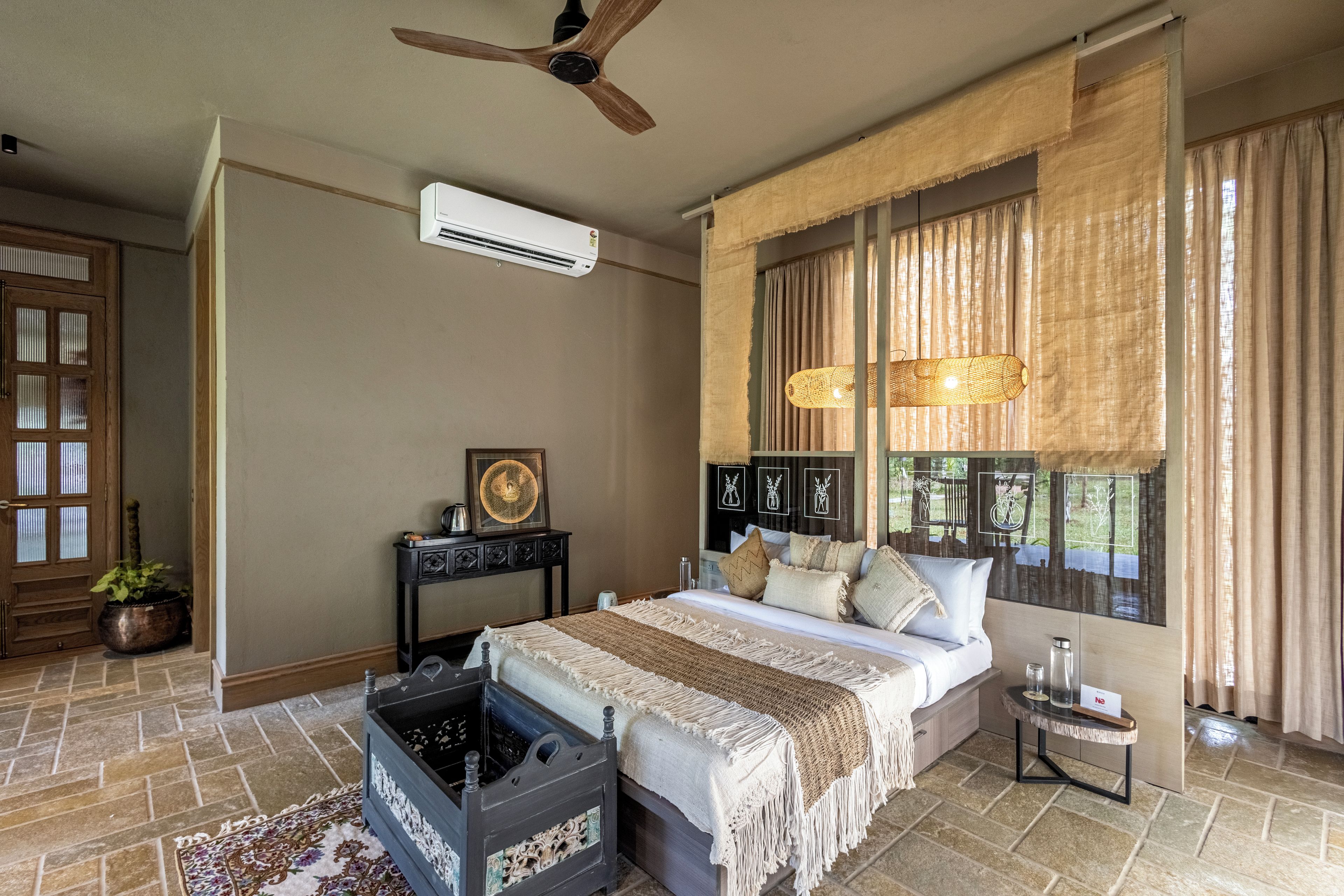 A bedroom with a bed, a ceiling fan, an air conditioning unit, a window with curtains, and a small table with a lamp.