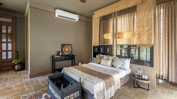 A bedroom with a bed, a ceiling fan, an air conditioning unit, a window with curtains, and a small table with a lamp.