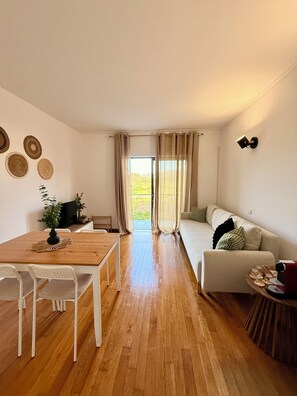 Habitació amb terra de fusta, un sofà, una taula de dinar amb cadires, un televisor i una finestra amb cortines.