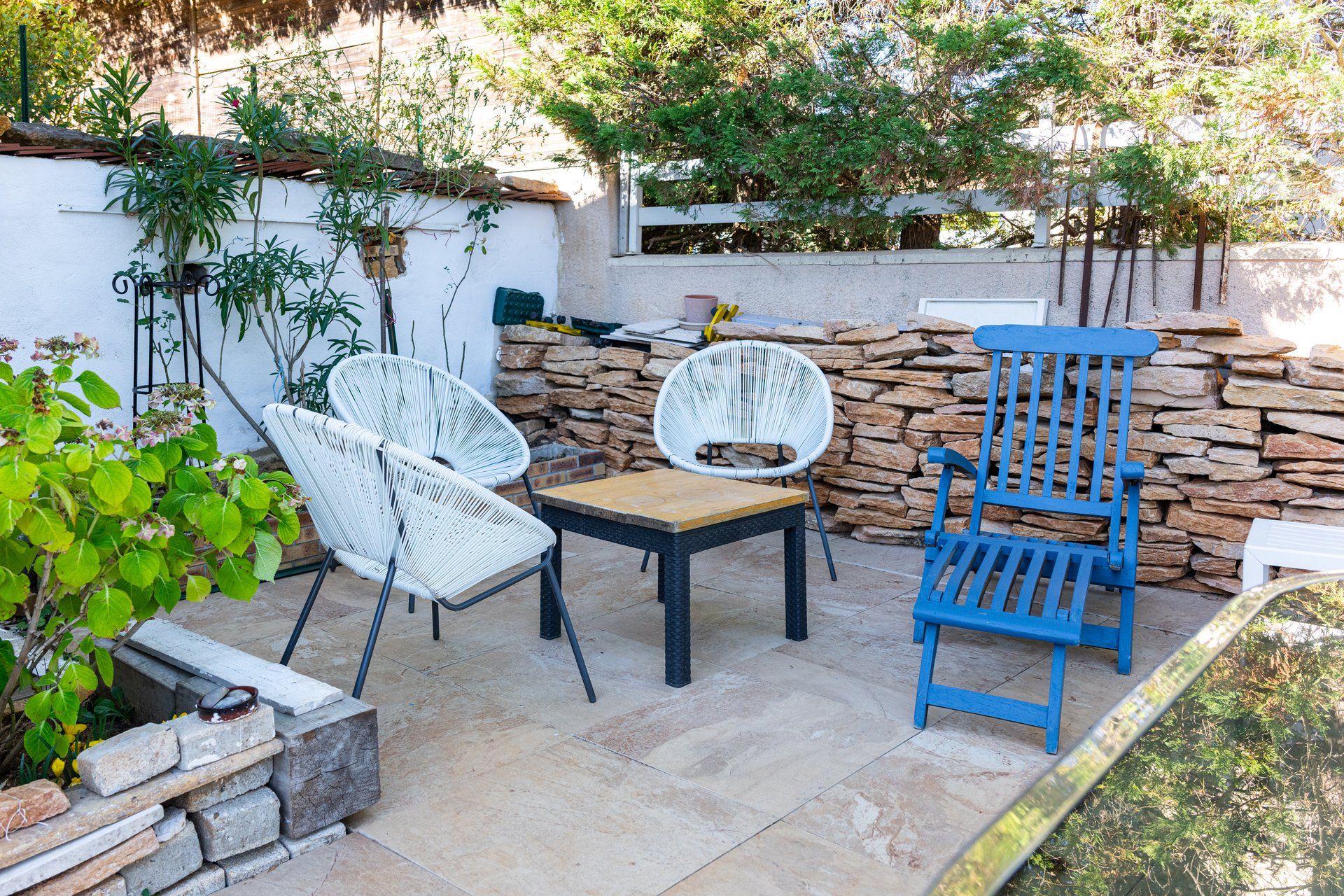 An outdoor seating area with stone walls, two white wicker chairs, a wooden table, and a blue wooden chair.