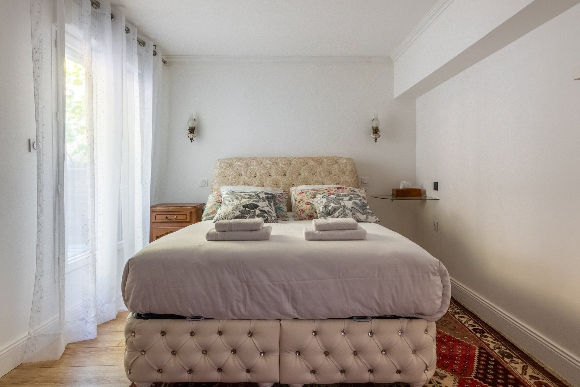 A neatly made bed with a tufted headboard, a bedside table, and a small shelf with a tissue box.