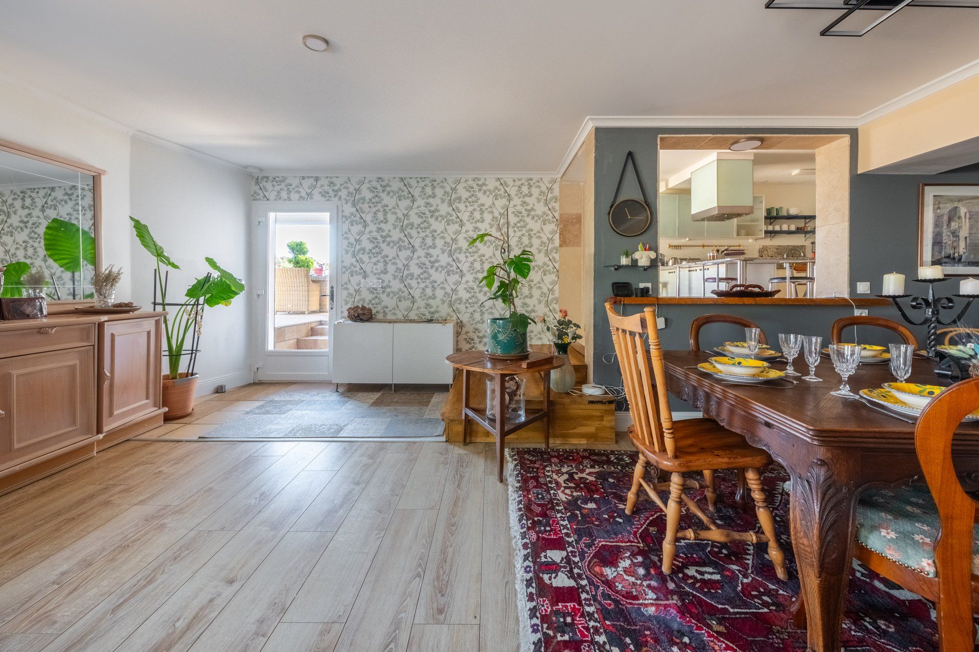 A dining area with a wooden table set for four, a patterned rug, and a view into the kitchen.
