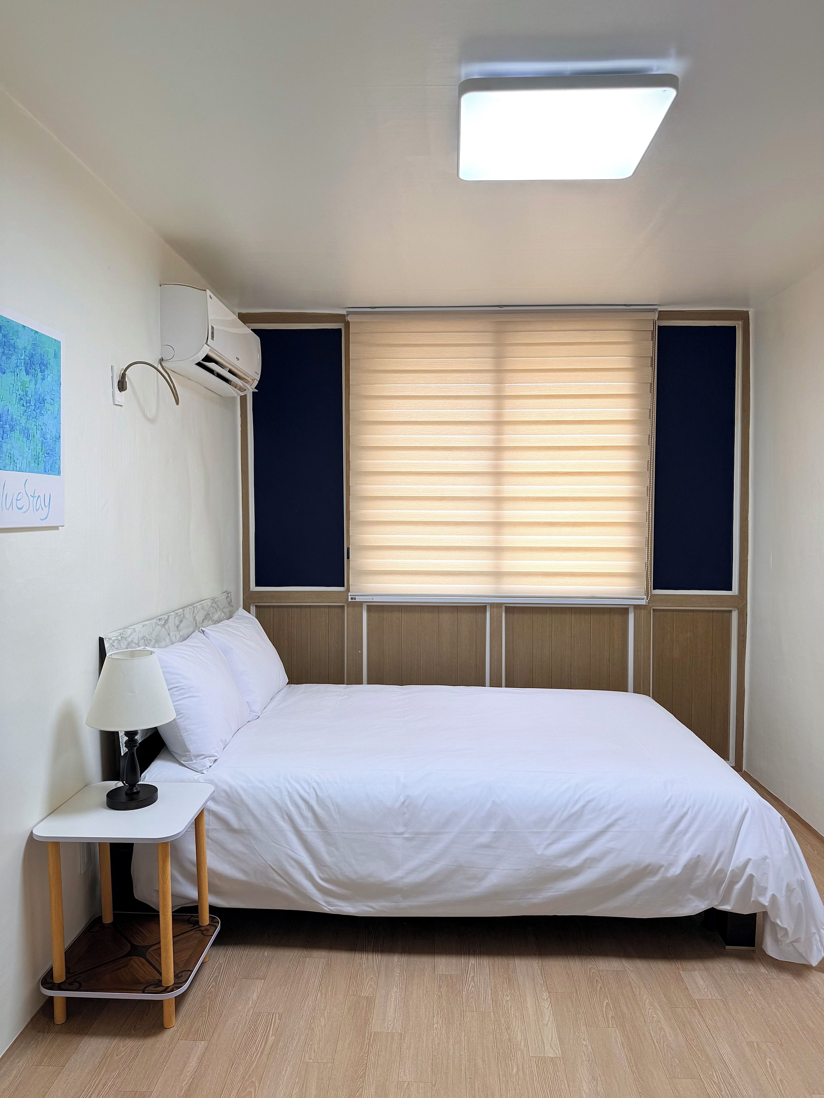 Un dormitorio con cama, mesita de noche, lámpara, unidad de aire acondicionado y ventana con persianas.