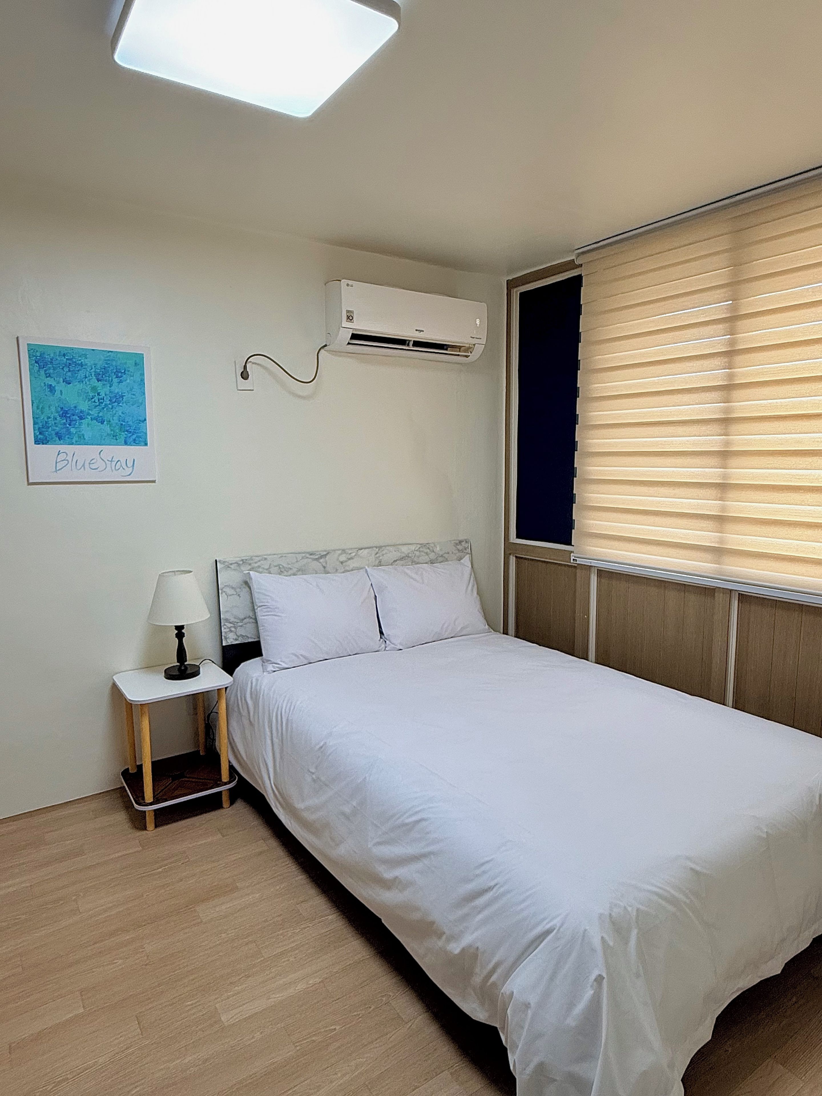 A bedroom with a bed, a bedside table with a lamp, an air conditioning unit, a window with blinds, and a wall-mounted artwork.