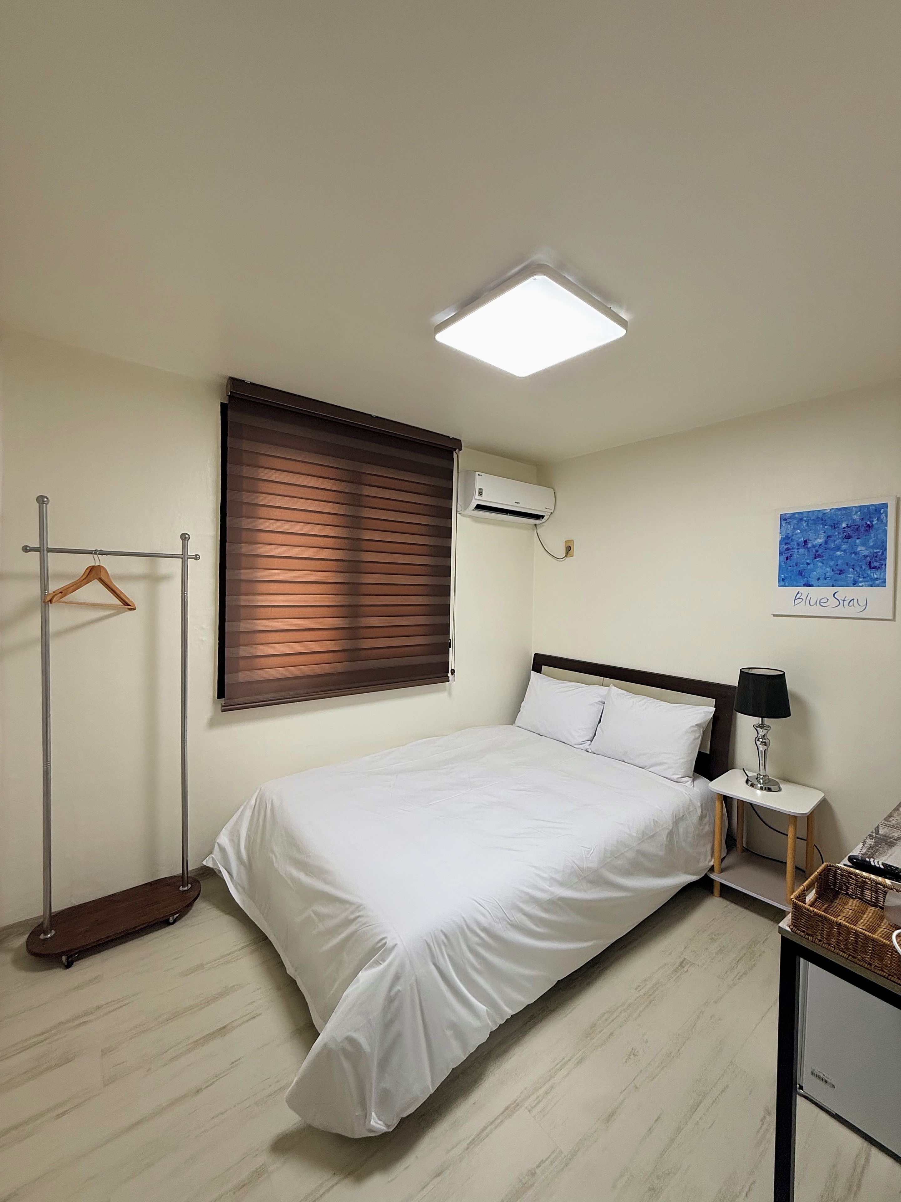 A hotel room with a bed, a bedside table with a lamp, a hanging coat rack, an air conditioning unit, and a window with blinds.