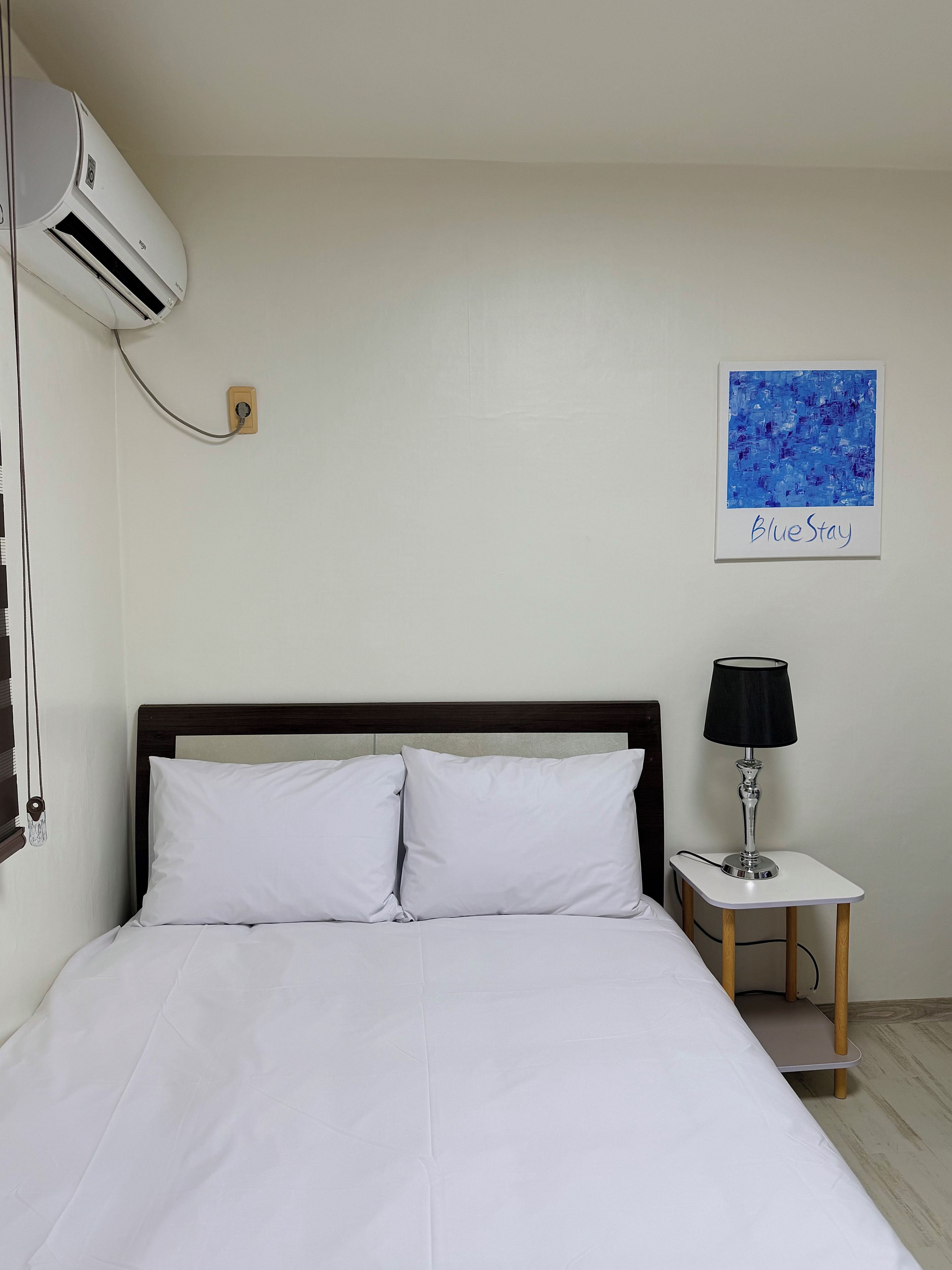 A hotel room with a bed, a bedside table with a lamp, an air conditioning unit, and a wall-mounted artwork.