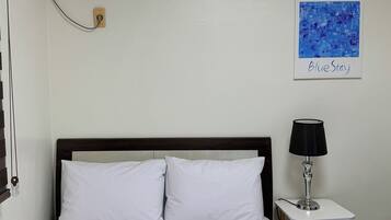 A hotel room with a bed, a bedside table with a lamp, an air conditioning unit, and a wall-mounted artwork.