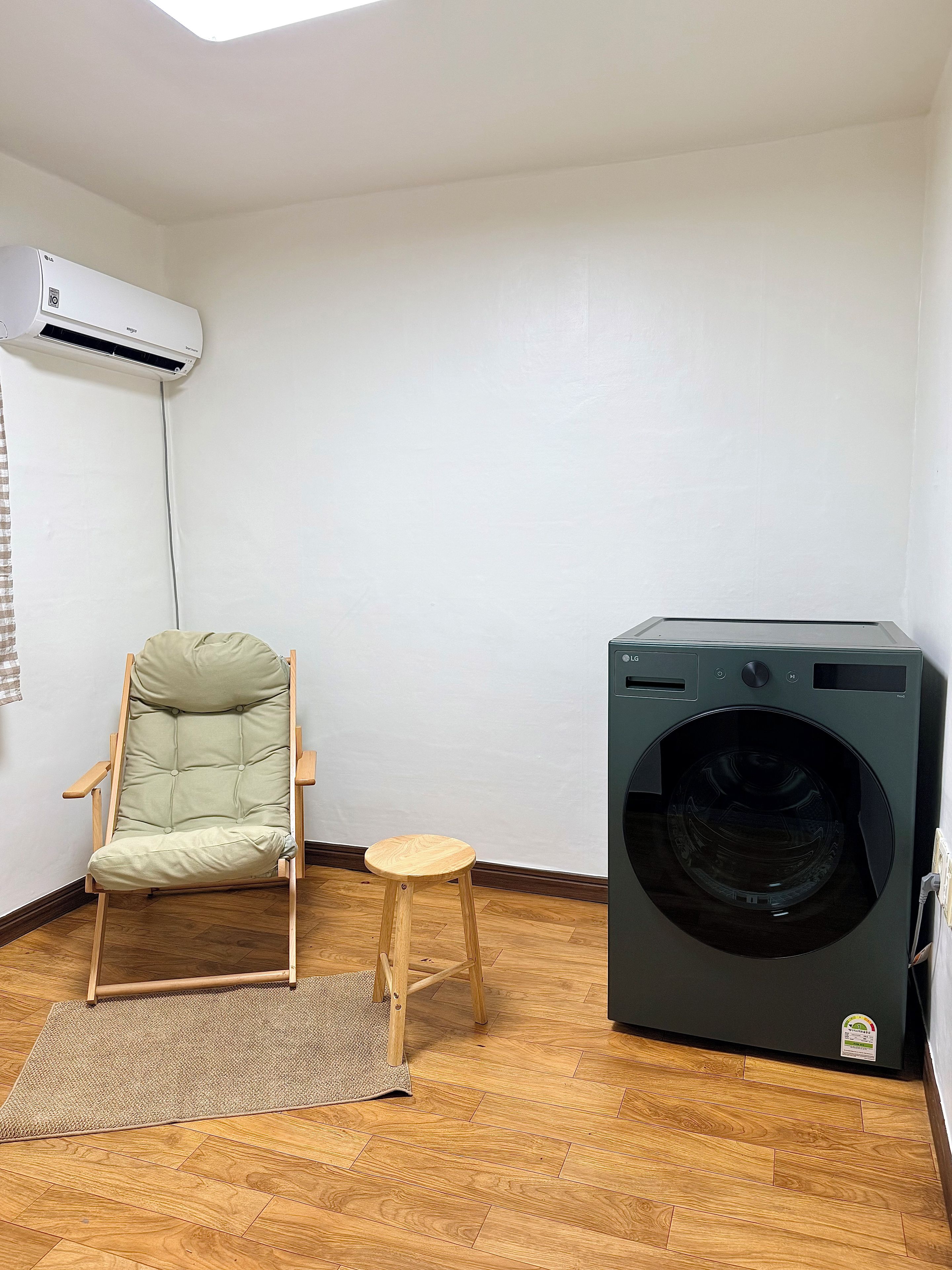 A room with a wooden floor, a green cushioned chair, a small stool, a washing machine, and an air conditioning unit.