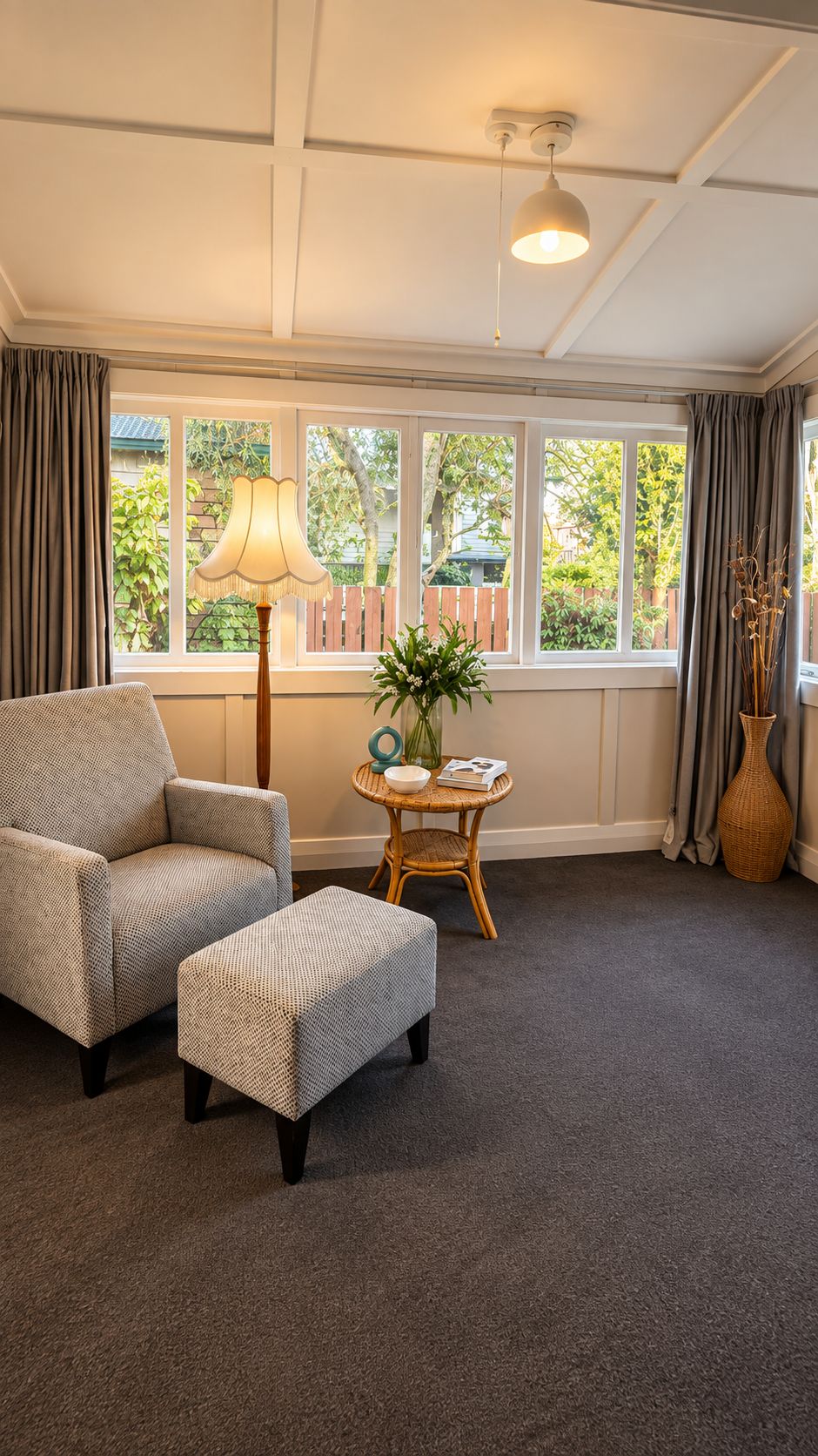 A room with a large window, a chair, an ottoman, a table with a lamp, and a vase with flowers.