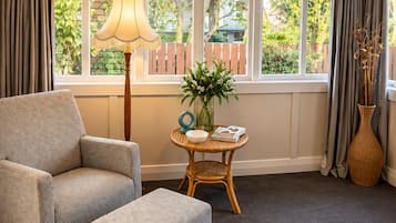 A room with a large window, a chair, an ottoman, a table with a lamp, and a vase with flowers.