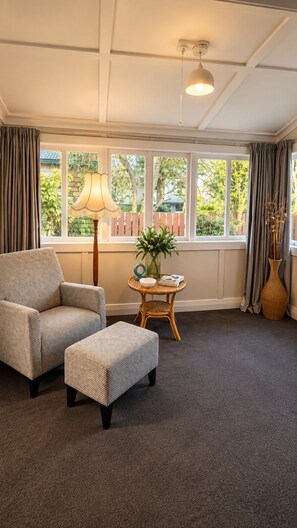Een kamer met een groot raam, een stoel, een voetenbankje, een tafel met een lamp en een vaas met bloemen.