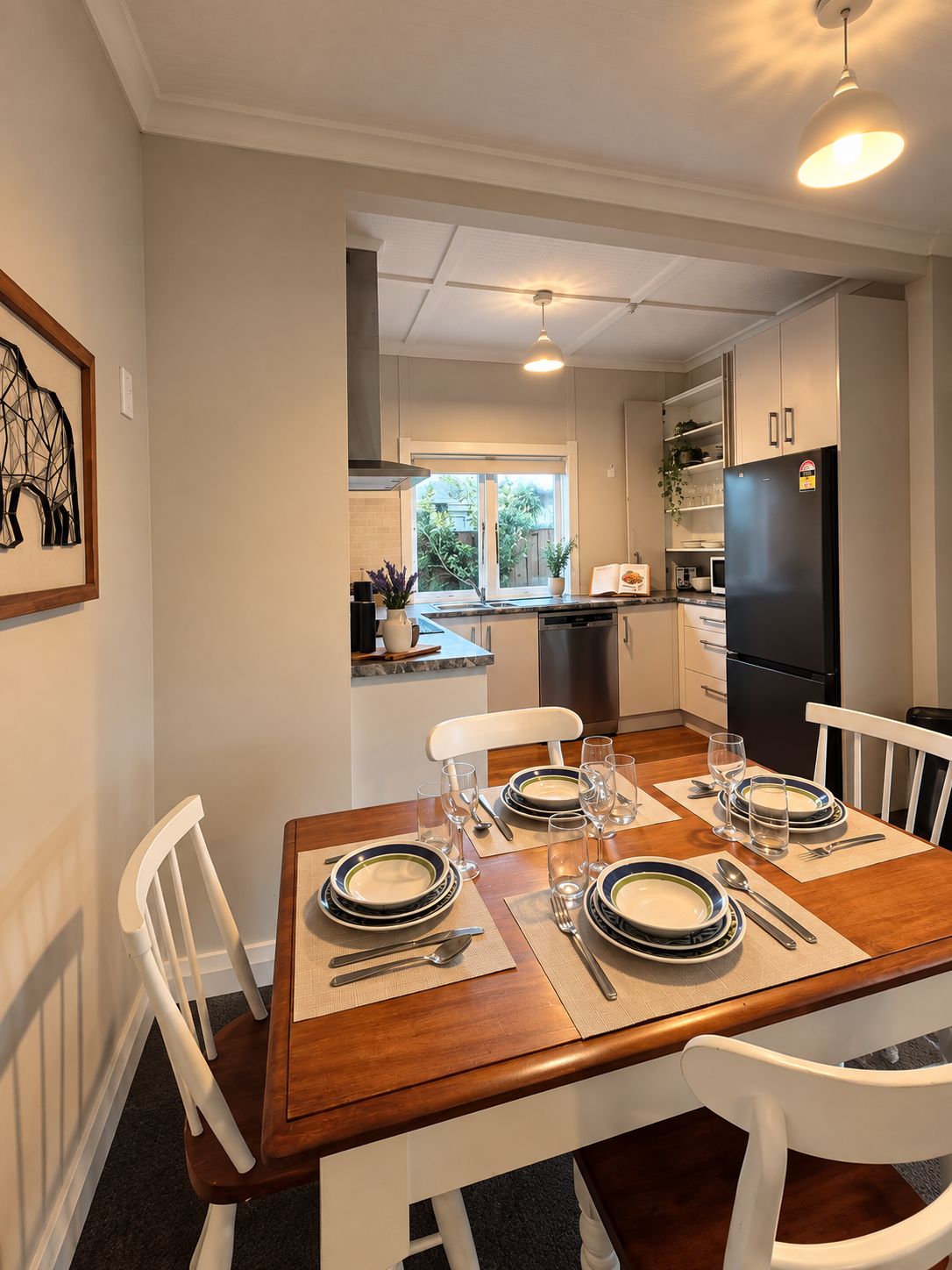 Un comedor con mesa de madera dispuesta para cuatro personas, sillas blancas y una cocina al fondo.