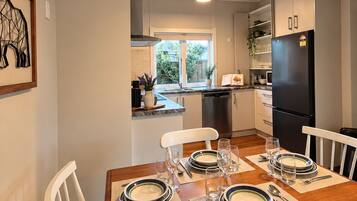 Un comedor con mesa de madera dispuesta para cuatro personas, sillas blancas y una cocina al fondo.