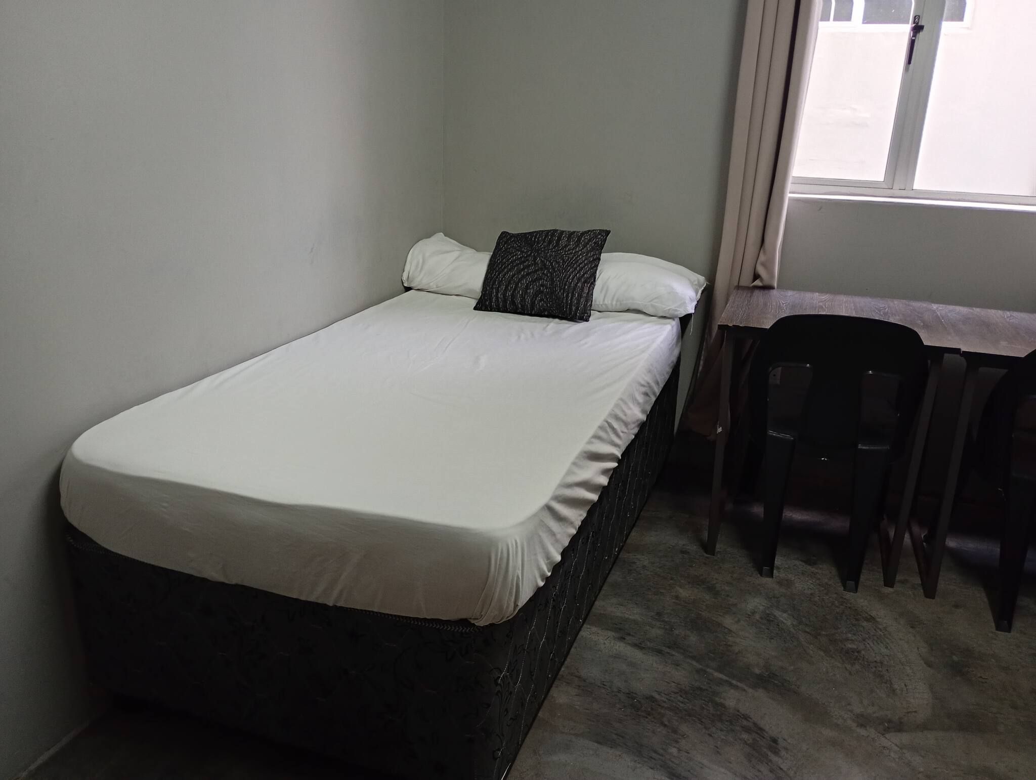 Een enkel bed met een witte matras en een donker hoofdbord, een tafeltje met een stoel, en een raam met gordijnen.