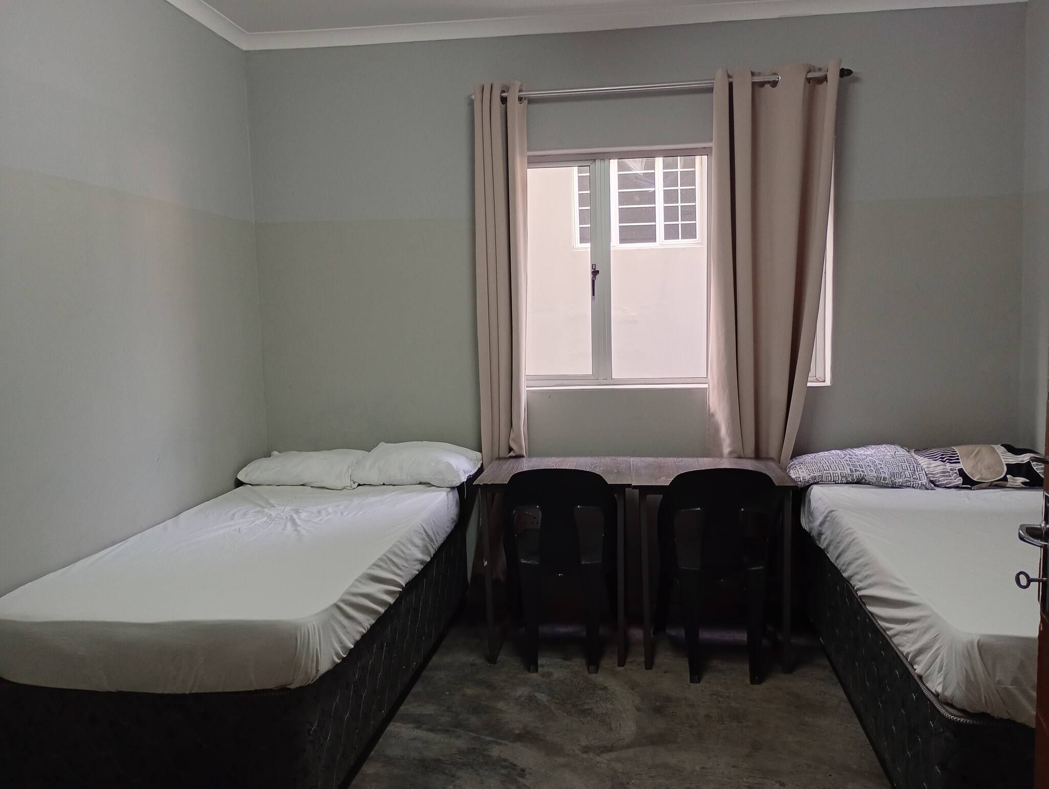 A small, single-room accommodation with two single beds, a small table, and a window with curtains.