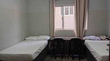 A small, single-room accommodation with two single beds, a small table, and a window with curtains.