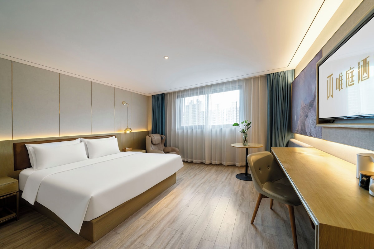 Habitación de hotel con una cama grande, un escritorio, una silla, una mesita con un jarrón y un ventanal con cortinas.