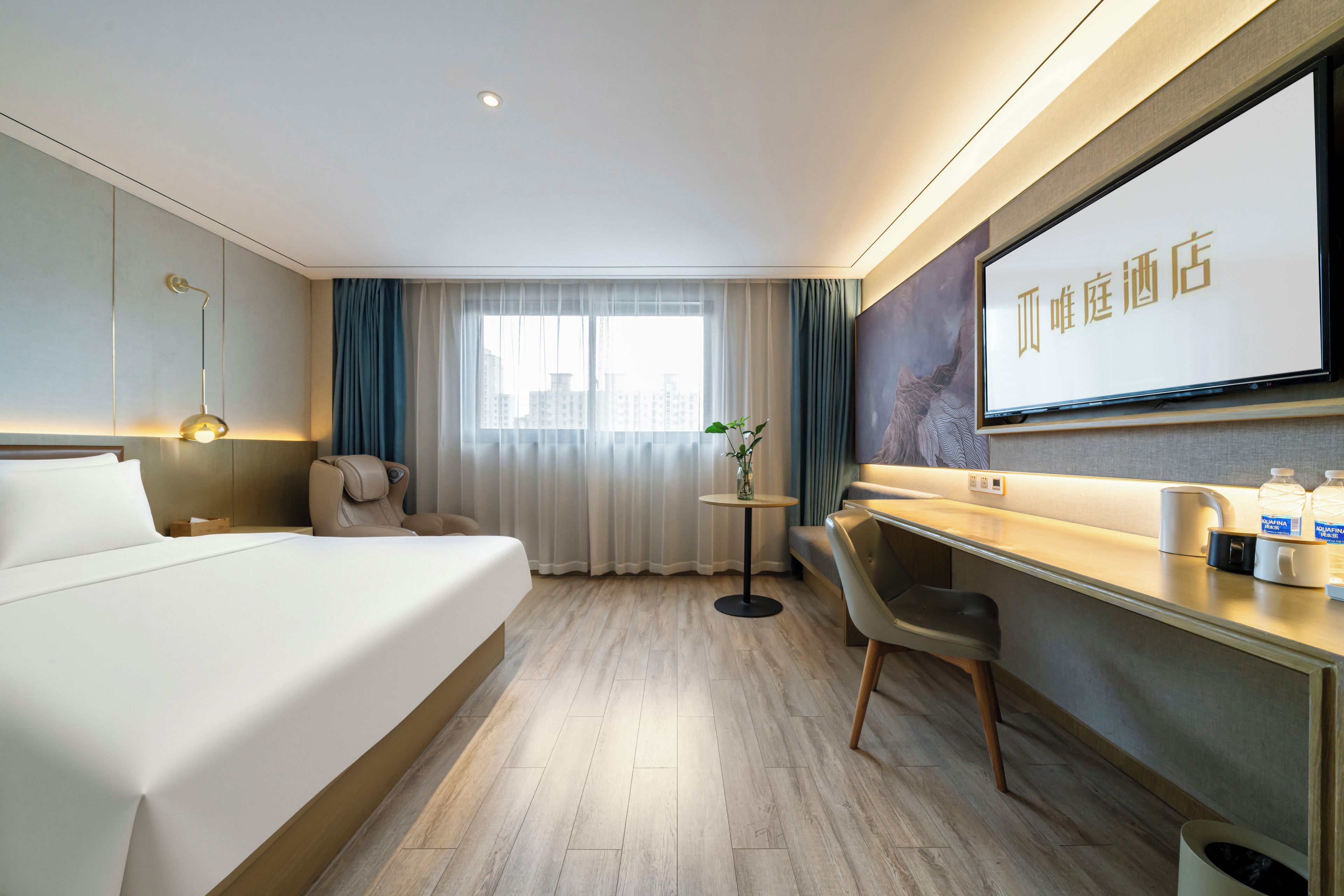 Habitación de hotel con cama, escritorio, silla, una pequeña mesa con un florero y una televisión grande mostrando texto.