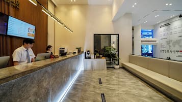 Een hotel receptie met een marmeren vloer, een receptiebalie, een digitaal scherm dat kamerprijzen toont en een muur met een kaart en bedrijfslogo's.