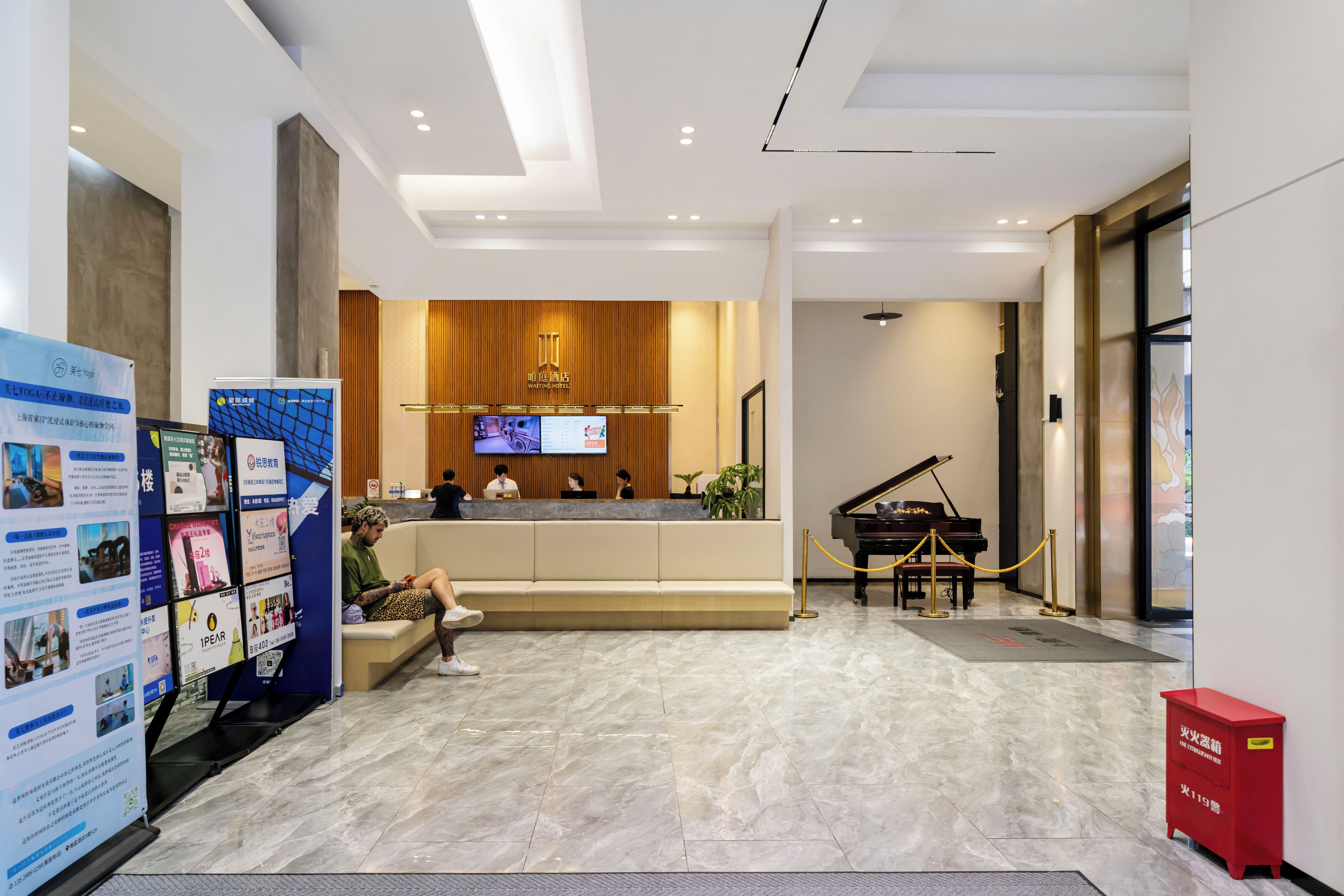 A hotel reception area with a reception desk, a television screen, a piano, and a red fire safety cabinet.