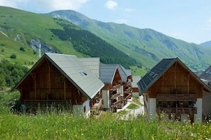 A row of wooden chalets with sloped roofs, set against a backdrop of green hills and mountains. - Sybelles Ski Area | Sleeps 12 | Slope-side Comfort (Saint Sorlin d'Arves)