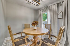 Un comedor con una mesa redonda de madera, cuatro sillas tapizadas, un jarrón con flores secas y una ventana con cortinas.