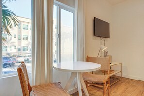 A room with a round table, two wicker chairs, a flat-screen TV mounted on the wall, and a view of the street outside. - Sunset Strolls & Seaside Fun: Santa Monica Condo! (Santa Monica)